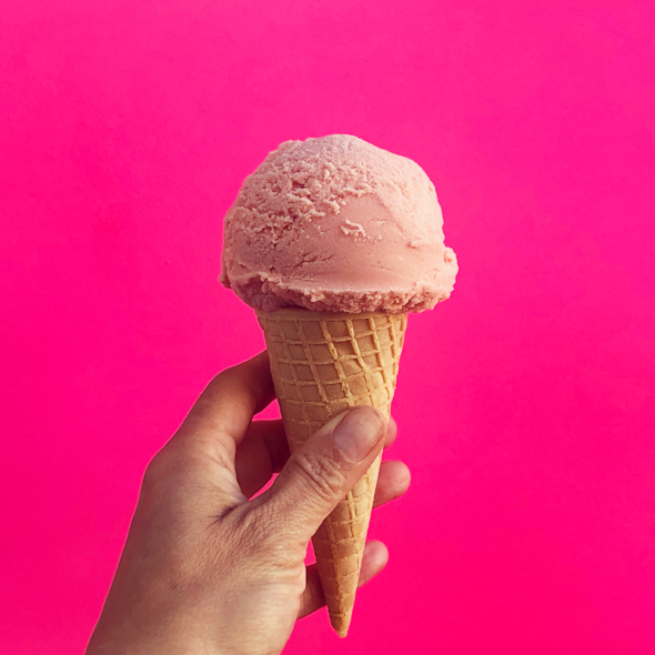 a pink scoop of ice cream in a waffle cone being held by a hand against a neon pink background